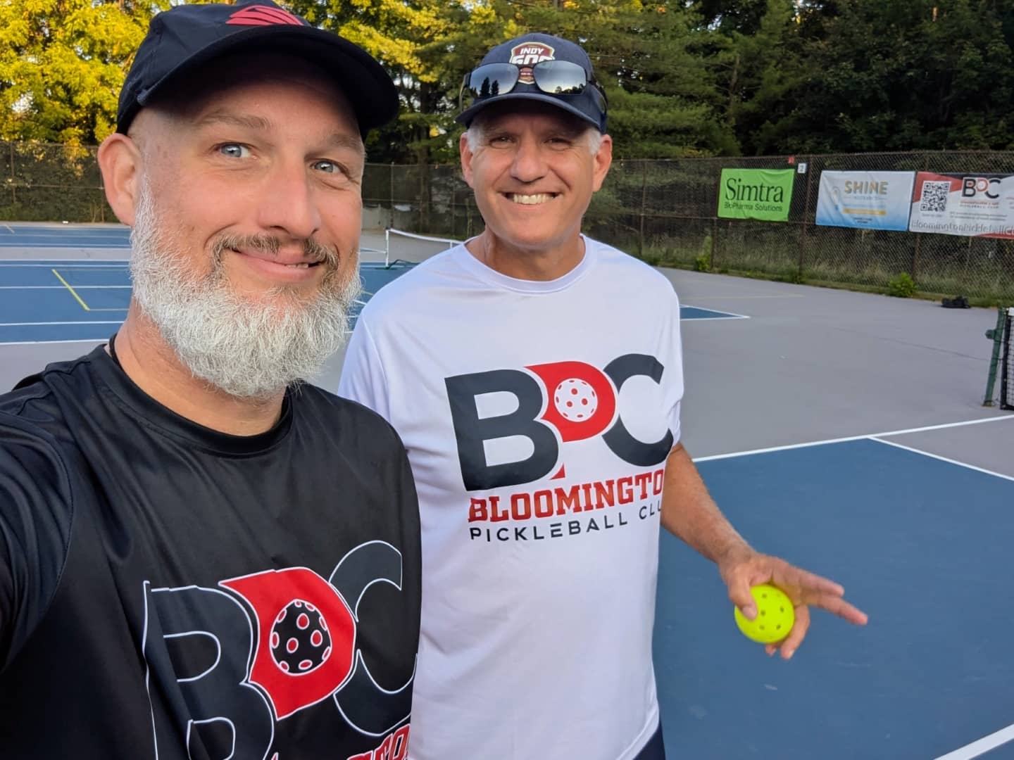 Dave Martin, a pickleball coach, poses with another player on an outdoor pickleball court in Bloomington, Indiana. Both wear Bloomington Pickleball Club shirts.