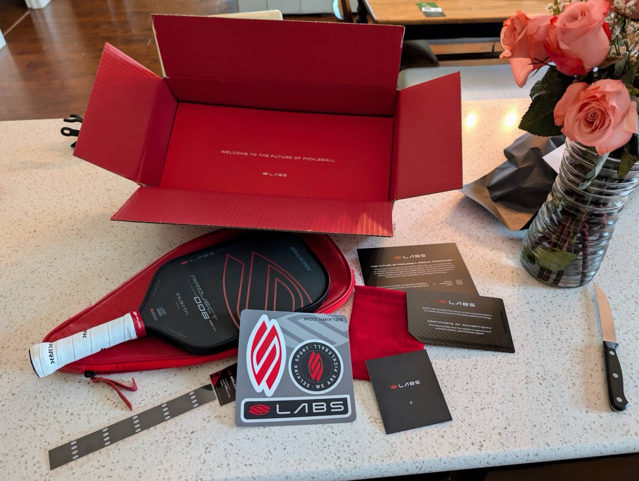 Selkirk Labs 008 pickleball paddle with red case, stickers, and packaging on a kitchen countertop with roses.