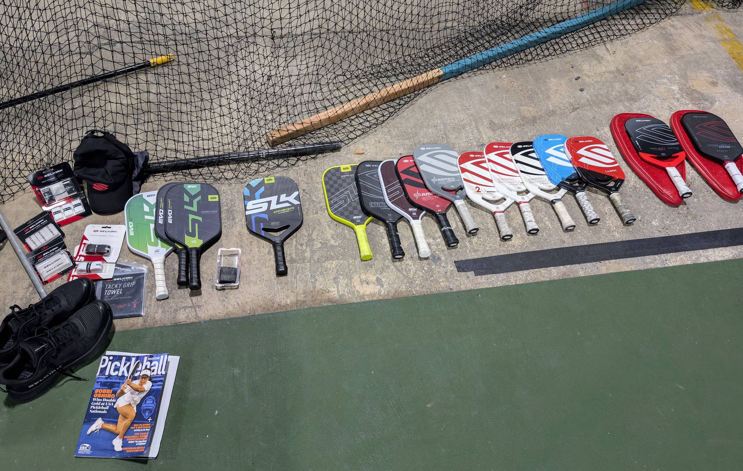 A collection of Selkirk pickleball paddles, accessories, and gear displayed on a court. Includes SLK Evo, Amped, Vanguard, and Labs series paddles along with grips, towels, and a Pickleball Magazine.